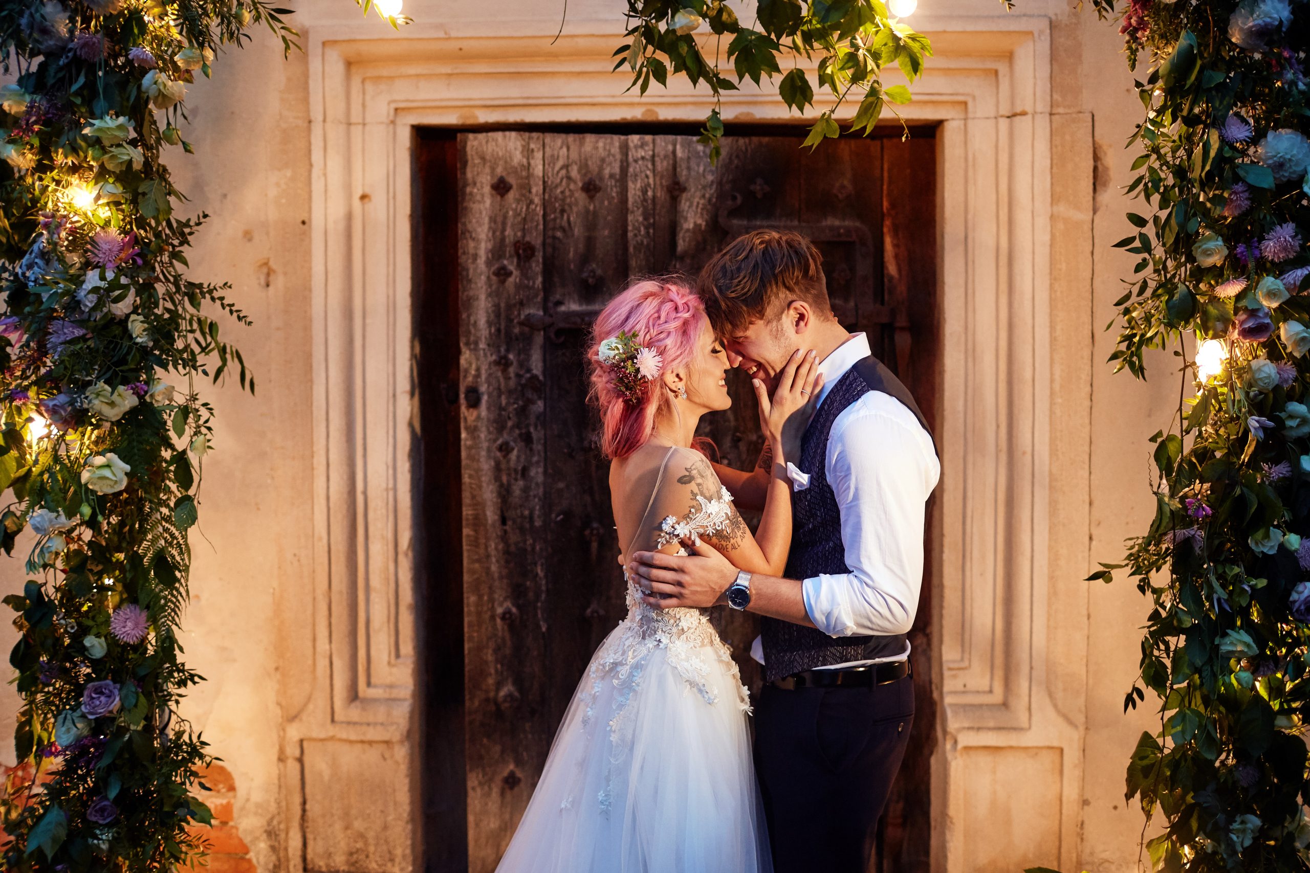 Groom and bride with pink hair stand before a door with flower g